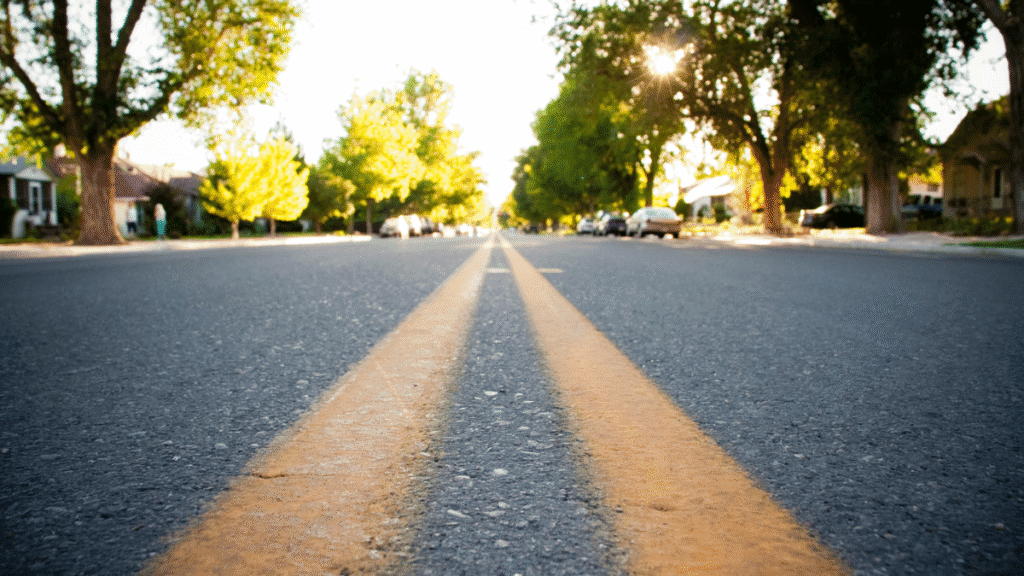 A low-angle view of a sunlit suburban street with clear double yellow lines. Green trees, houses, and parked cars line the peaceful road.
